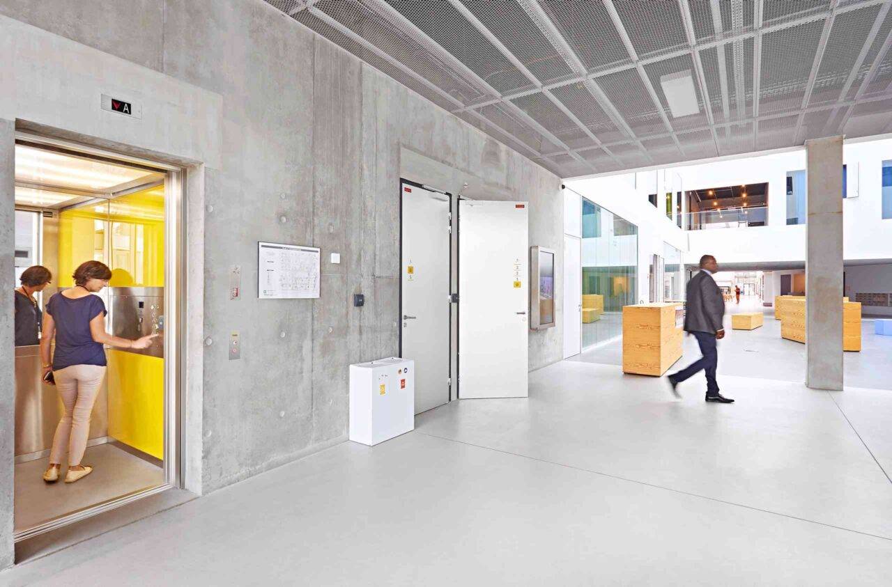 A modern indoor space with yellow and wooden accents, featuring an elevator, benches, and large windows.
