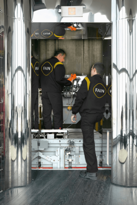 Three technicians in black uniforms marked "FAIN" working inside an elevator shaft.